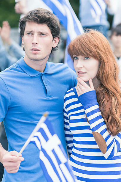 Amid chants and raised banners, a Greek couple joins the protest, their Greek flags waving as a sign of unity and defiance against mass home auctions
