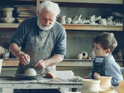 A grandfather teaching a young boy to work with clay at a pottery wheel, symbolising the tradition of learning through patient guidance and shared experience