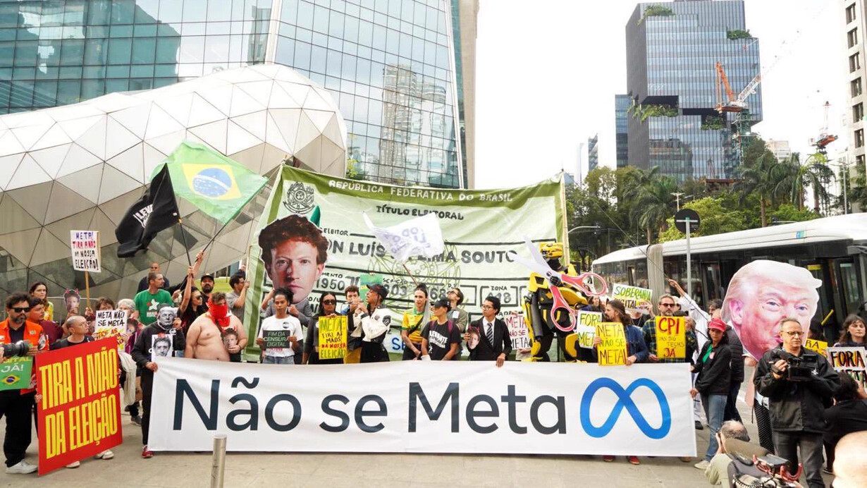 Manifestantes se reúnem em São Paulo diante da sede da Meta, empresa responsável por plataformas como Facebook, Instagram e WhatsApp, para protestar contra a possível interferência das redes sociais no processo eleitoral brasileiro. A faixa central resume a reivindicação: “Não se Meta”, em um jogo de palavras que transforma o nome da companhia em uma denúncia política. Entre bandeiras, cartazes e imagens de figuras públicas, outras mensagens pedem: “Tira a mão da eleição”. A manifestação expressa a preocupação com o poder das grandes plataformas de influenciar o debate público, determinar a visibilidade de discursos e interferir na maneira como informações e opiniões circulam. Em uma sociedade na qual a disputa política também se trava por meio de algoritmos, memes e campanhas digitais, a fotografia evoca uma questão central: quem controla os mecanismos que ajudam a formar a opinião pública e até que ponto esse poder pode afetar a democracia? A fotografia corresponde ao protesto realizado em 19 de outubro de 2026, diante da sede da Meta, na Avenida Faria Lima, em São Paulo