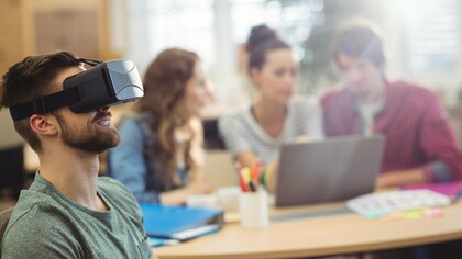 Um homem utiliza um equipamento de realidade virtual enquanto outras pessoas trabalham ao fundo. As novas tecnologias transformam o ambiente profissional, mas não eliminam a importância das relações humanas nas organizações. A evolução tecnológica avança rapidamente, desafiando profissionais e empresas a se adaptarem continuamente. O desafio permanece o mesmo: equilibrar inovação, desenvolvimento humano e construção de carreiras com propósito