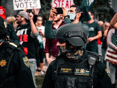Amidst a crowded protest during the day, a man in a black police uniform stands, representing the increasing polarization and the struggle for control in today's fractured social landscape