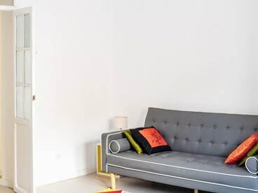 Um sofá cinza se destaca diante da parede clara, enquanto um tapete colorido e elementos decorativos acrescentam personalidade à sala. Como observa o texto, móveis, cores e objetos também podem revelar a relação de seus moradores com a estética e com a imagem que desejam projetar