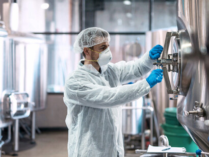 A person wearing protective gear (hairnet, mask, gloves, and a lab coat) working with a large stainless steel industrial tank