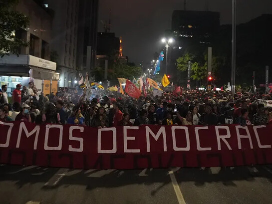 Uma multidão ergue a faixa “somos democracia” durante protesto popular. A disputa entre participação democrática e projetos de poder autoritários está no centro das tensões políticas contemporâneas