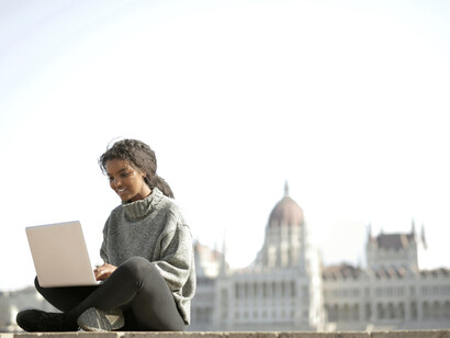 In a sunny European plaza, a woman in a gray long-sleeve shirt types on her laptop, blending work with travel