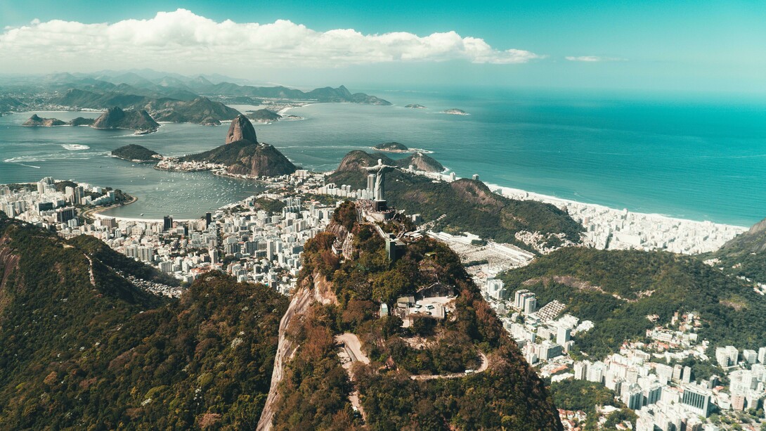 Vista aérea do Rio de Janeiro. Muito antes da chegada dos portugueses, a região da Baía de Guanabara já era habitada por povos indígenas, especialmente do tronco Tupi-Guarani. Os Tupinambás e os Tamoios viviam da pesca, da caça, do cultivo de mandioca e da coleta de frutos, em um sistema de vida baseado na relação com a natureza. Esses povos chamavam a região de “Guanabara”, que significa “seio do mar” em tupi, em referência à entrada da baía que hoje abriga a cidade. Era uma área estratégica por suas condições naturais de abrigo e abundância de recursos