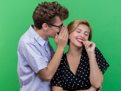 A young, beautiful couple stands against a green wall as the man whispers a secret to his smiling girlfriend.