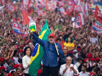 Lula aparece no centro da imagem, diante de uma multidão de apoiadores, segurando um exemplar da Constituição brasileira enquanto uma grande bandeira nacional se destaca atrás dele. Ao redor, predominam bandeiras, camisetas e bonés vermelhos, compondo o cenário de uma mobilização política de grandes proporções. O gesto de erguer a Constituição ganha significado especial em um contexto no qual a democracia brasileira enfrenta disputas sobre suas instituições, seus limites e sua capacidade de garantir direitos. No artigo, Boaventura de Sousa Santos chama a atenção para o desafio de preservar a democracia para além das urnas, fortalecendo a participação popular e a resistência às forças autoritárias