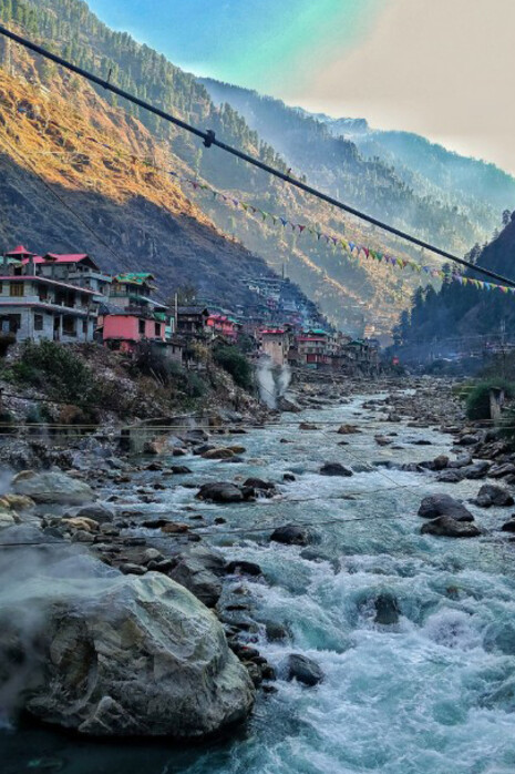 A river flows gracefully through the lush valley of Manikaran, Himachal Pradesh, surrounded by majestic Himalayan mountains, India