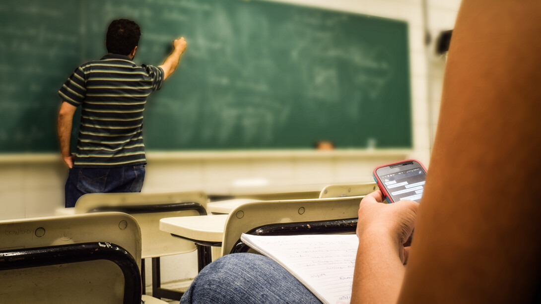 Professor em sala de aula ensinando uma matéria na lousa e a visão do lugar do aluno, que se distrai ao usar o celular