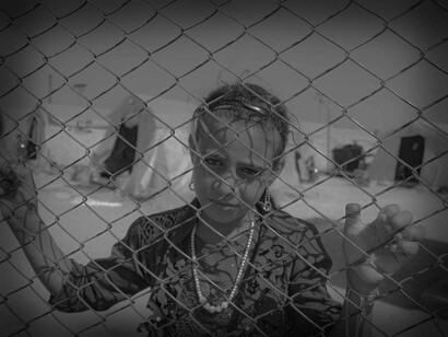 A little girl child looks at the camera from behind the fence of a refugee camp in Syria