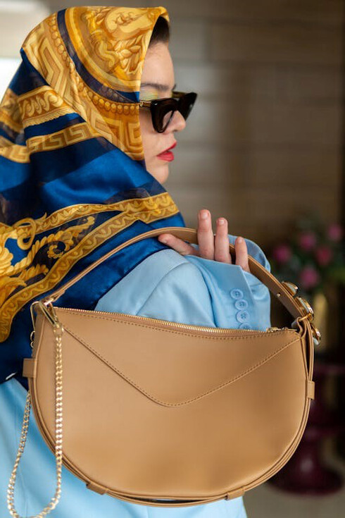 A sophisticated woman exudes elegance, seated in a light blue suit complemented by a refined headscarf, sleek sunglasses, and timeless black heels