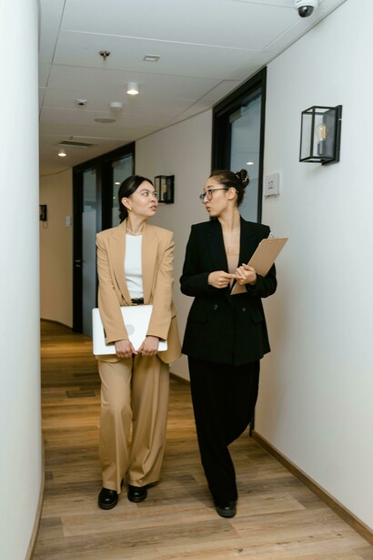 Two women in business casual attire are having a conversation as they walk along a corridor