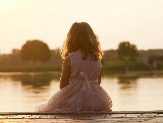 A young girl sitting alone beside the water at sunset, representing the loneliness that can begin with the fear of being left