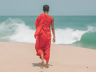 On the seashore, a figure clad in Buddhist attire gazes thoughtfully at the horizon