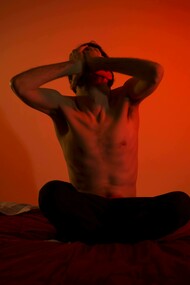 A topless man sits alone on his bed, his posture reflecting anguish and anxiety in a quiet, dimly lit bedroom