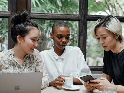 Happy multiethnic women chatting and using gadgets in a cafeteria, representing connection through apps and social media in everyday life