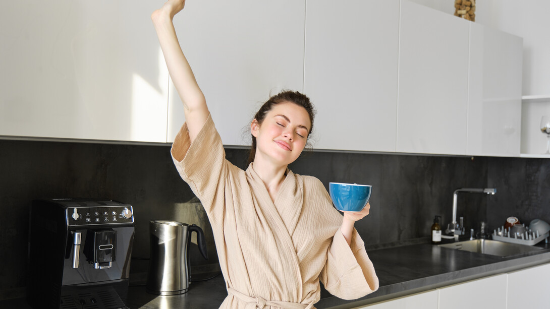 Brunette woman dancing and stretching in the kitchen while drinking coffee