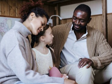 Pais e filha compartilham um momento de atenção e afeto no sofá da sala. O artigo destaca a importância da proximidade familiar na transmissão de valores e ensinamentos entre gerações