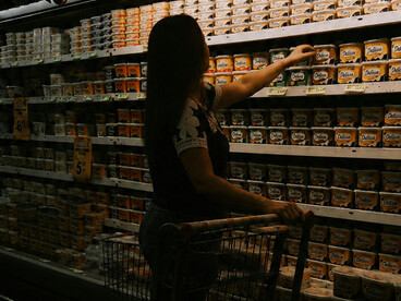 A silhouetted woman pushes a shopping cart through a dark grocery aisle lined with illuminated shelves of product