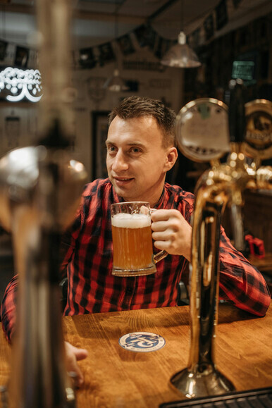 A man in a checkered long-sleeve shirt enjoys tasting beer, glass in hand