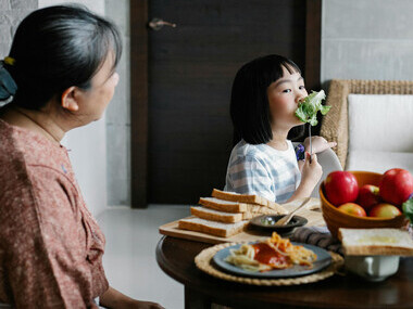 A grandmother sitting at the table enjoying a healthy salad with her Asian grandchildren, as a happy little girl looks at her