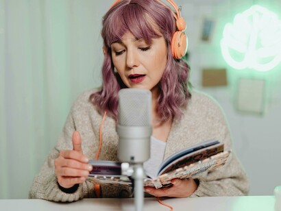 A podcast host reads from a book during a casual indoor recording session, wearing headphones and speaking into a microphone