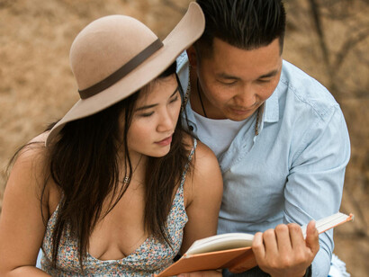 Mujer con vestido floral y con un sombrero marrón junto a un hombre con camisa de vestir celeste, leyendo un libro