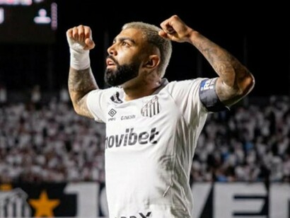 Gabigol ergue os braços e olha para o alto, vestindo a camisa do Santos e a braçadeira de capitão, enquanto as arquibancadas aparecem desfocadas ao fundo. A imagem destaca a postura confiante do atacante em um ambiente marcado pela presença da torcida. Depois de passagens por diferentes clubes e de temporadas de altos e baixos, seu retorno ao Santos abre um novo capítulo, no qual precisará corresponder às expectativas criadas em torno de seu nome