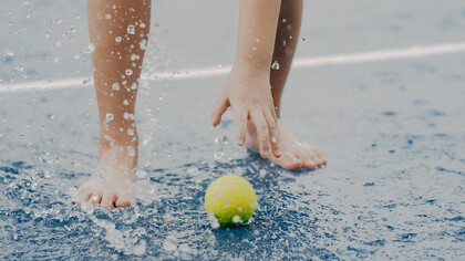Criança busca bola de tênis em quadra molhada de chuva