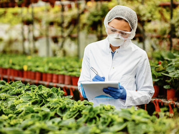 In a modern greenhouse, a female scientist examines potted flowers and records her observations, showcasing advances in plant science and biotechnology innovation