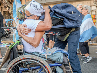 Momento emotivo durante una manifestación donde dos personas se abrazan cálidamente, una de ellas sentada en una silla de ruedas con banderas de Argentina de fondo