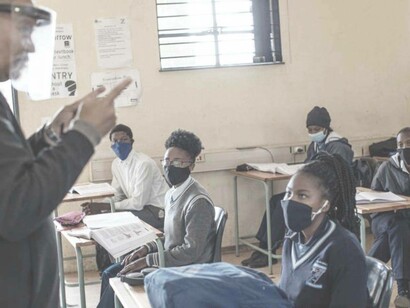 A teacher in a classroom in a Covid-19 setting is wearing a face cover guard while the youth of South Africa are wearing masks and listening attentively