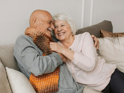 An elderly man in a gray sweater hugs the woman beside him, symbolizing emotional connection and overall elderly wellness