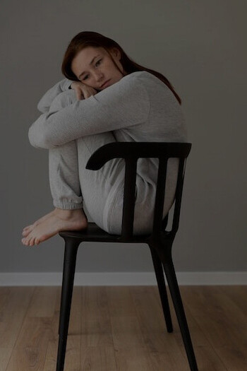 A woman sits quietly on a chair, her posture showing signs of stress and anxiety