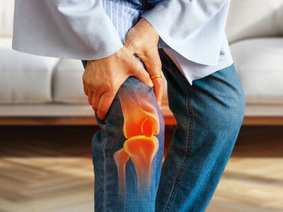 An elderly woman gently holds her knee in her living room, illustrating arthritis-related joint pain and knee injury