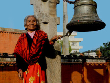 In Bhaktapur, Nepal, a senior Indian woman smiles warmly beside an old temple bell