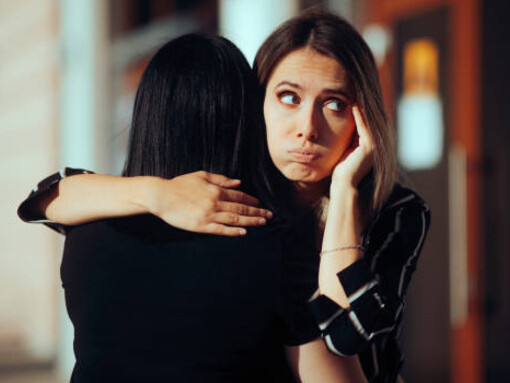 A woman warmly hugs a friend who smirks behind her back