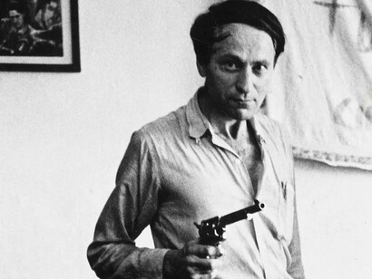 Jonas Mekas wielding a gun while smiling with his eyes