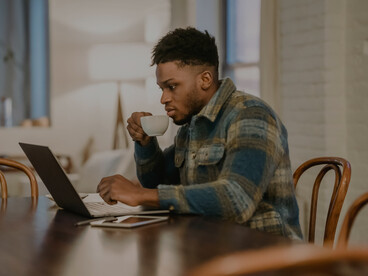 A focused Black man working on a laptop and sipping coffee in a café