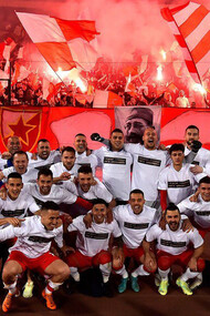 Jogadores do time Estre Vermelha, seleção de fuebol da República da Sérvia, juntos em foto enquanto a torcida ultra ao fundo comemora com a tradicicional queima de artifícios pirotécnicos