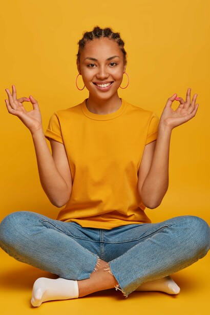A woman sitting on the floor and smiling in a meditative pose, her thumb and index finger gently touching
