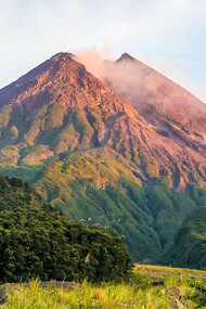 The serene beauty of Mount Merapi in Indonesia stands in stark contrast to its powerful history, with lush greenery enveloping its slopes and the clear blue skies above
