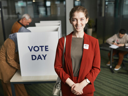 A woman in a red coat standing in front a voting booth while other people are casting their vote behind her