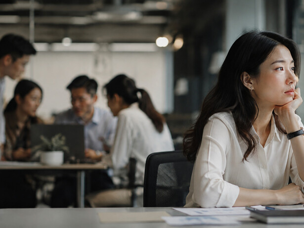 A woman sitting apart from her coworkers in a busy office, representing the emotional distance that can exist within a workplace