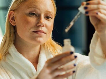 A woman in a white robe holding a dropper in front of a mirror, representing the gap between promising ingredients and what the skin can actually absorb