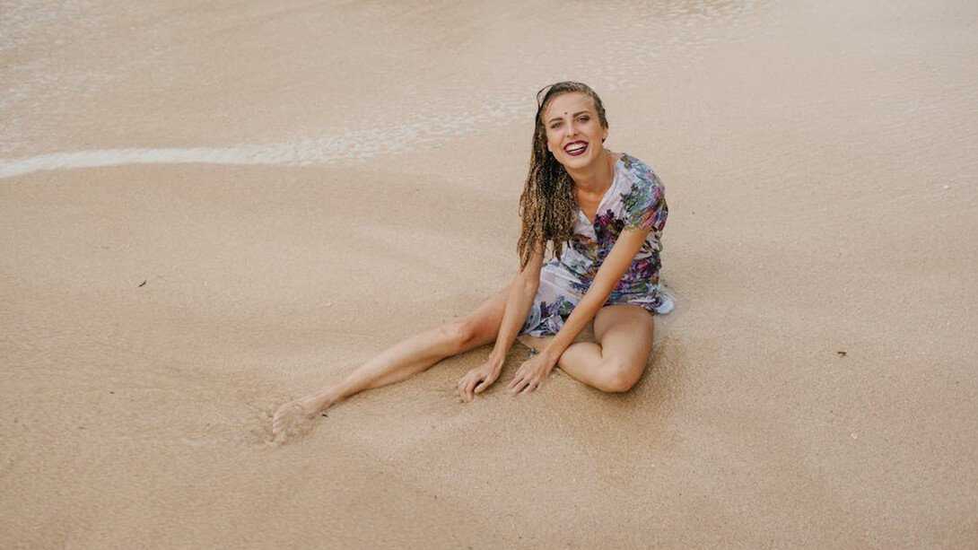 A cheerful and radiant woman sits on the sandy beach, wearing a smile that brightens the surroundings