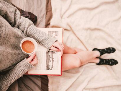 Mujer acostada en cama, lee un libro junto con una taza de café