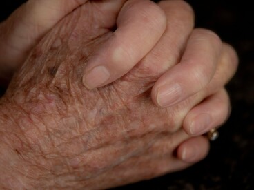 Em um enquadramento fechado, mãos envelhecidas aparecem unidas, com destaque para as rugas e as dobras da pele. Cada marca evoca a passagem dos anos e a trajetória de quem viveu, sentiu e construiu vínculos. O corpo se transforma com o tempo, mas essas mudanças não diminuem o valor nem a história de uma pessoa