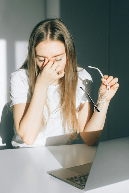 A young woman pauses in a bright office, holding her glasses as she struggles with digital eye strain and exhaustion brought on by extended hours in front of screens